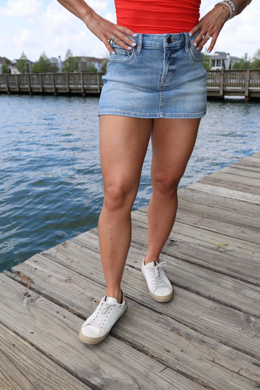 Person wearing a denim skirt and white sneakers on a wooden dock by water