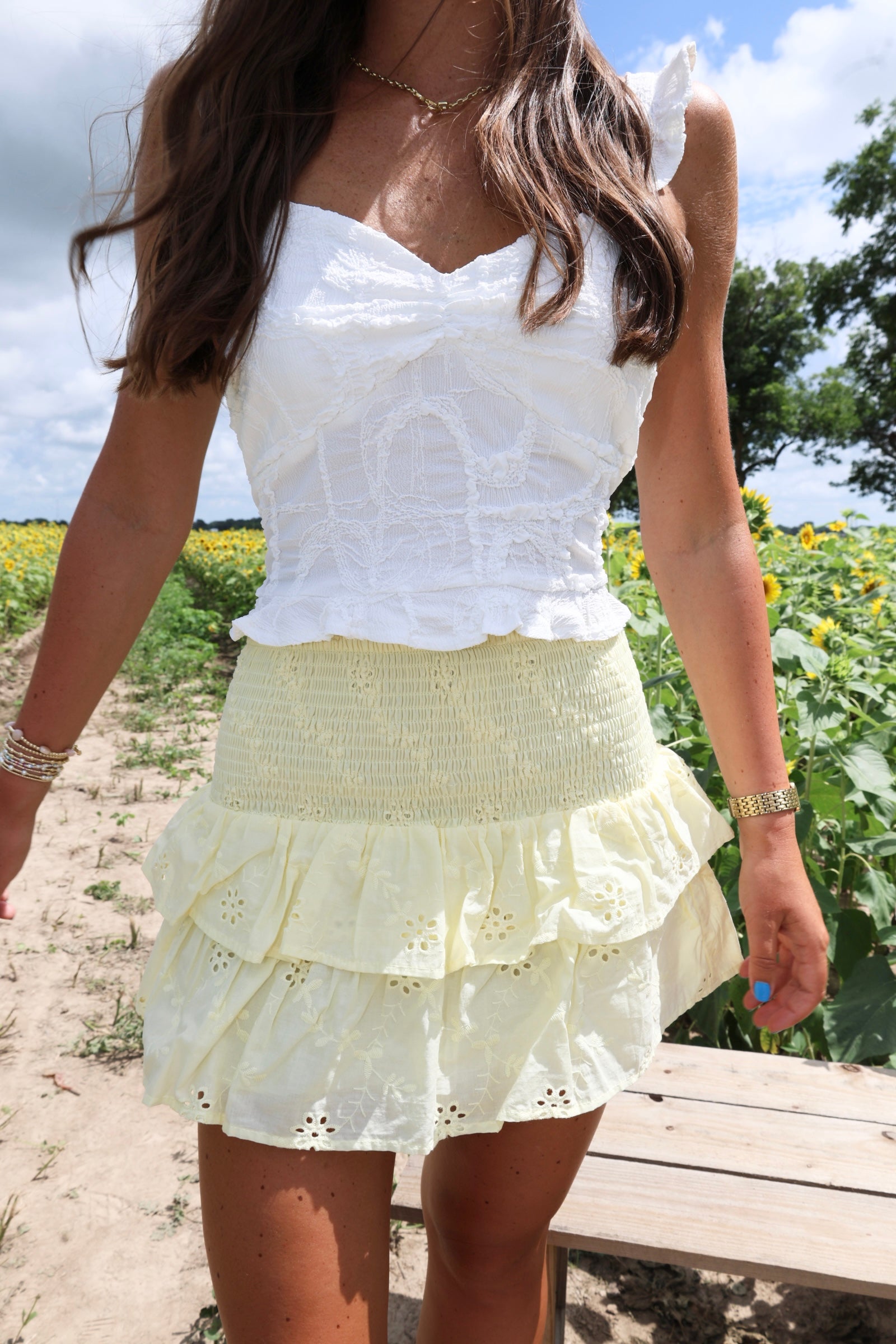Woman wearing a white top and yellow skirt standing in a field with sunflowers.
