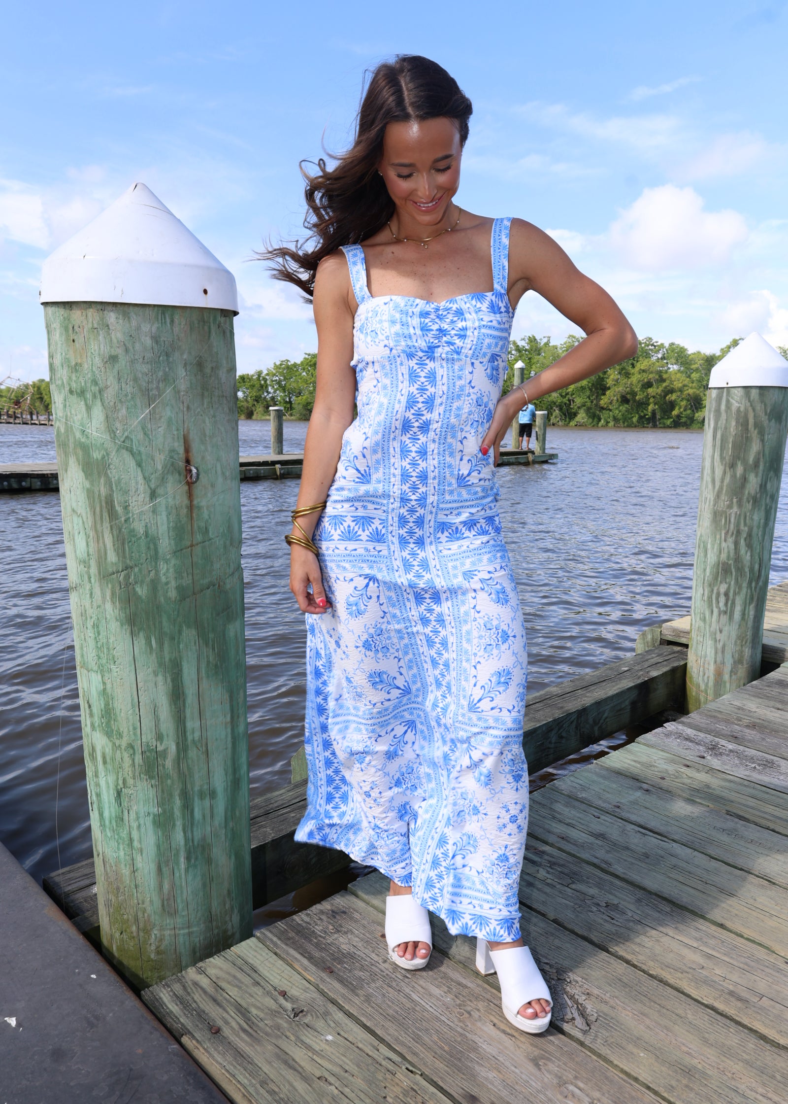 Woman in a blue and white patterned dress standing on a wooden dock by water.
