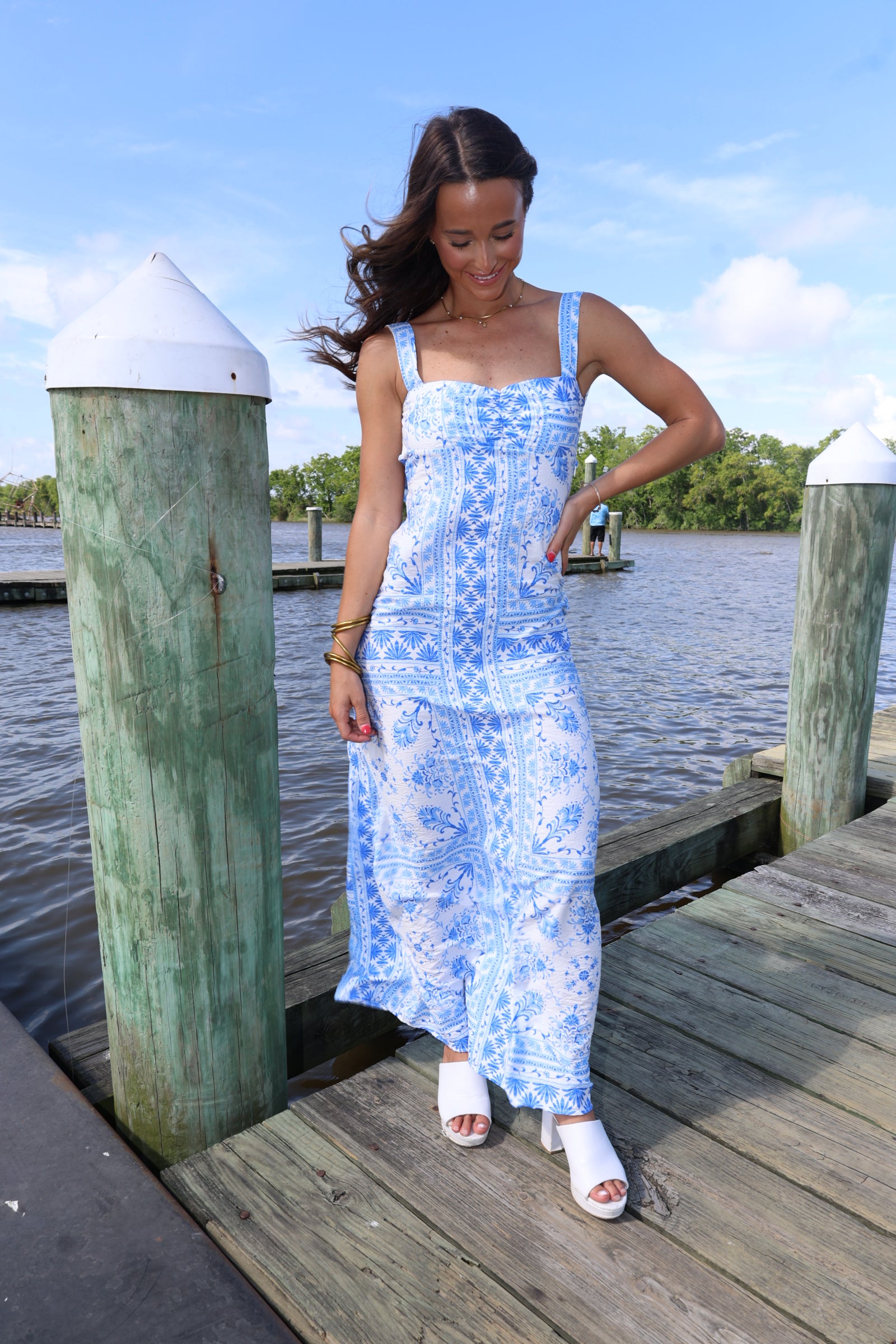 Woman in a blue and white patterned dress standing on a wooden dock by water.