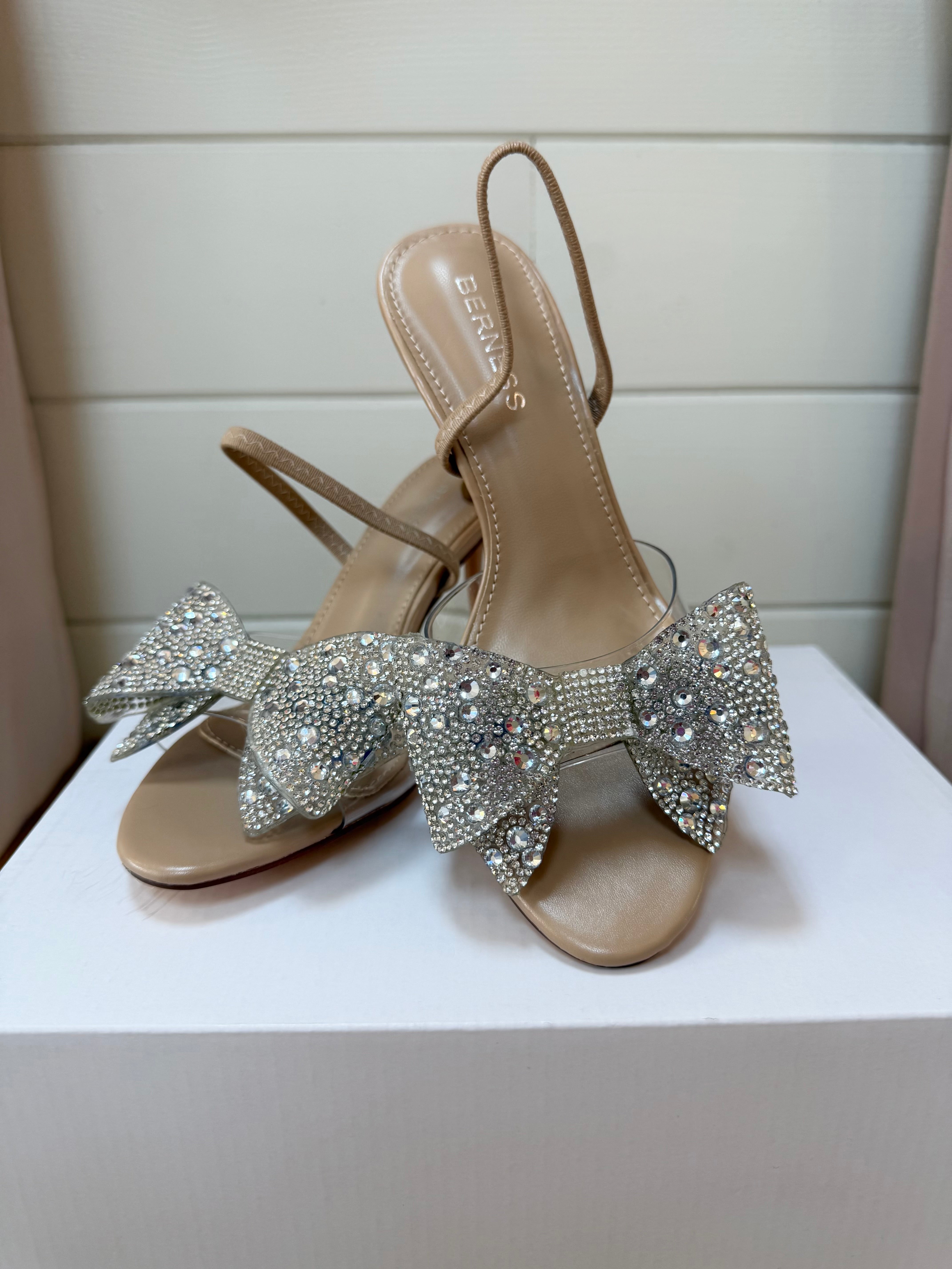 Beige high-heeled sandals with large crystal bows on a white surface.