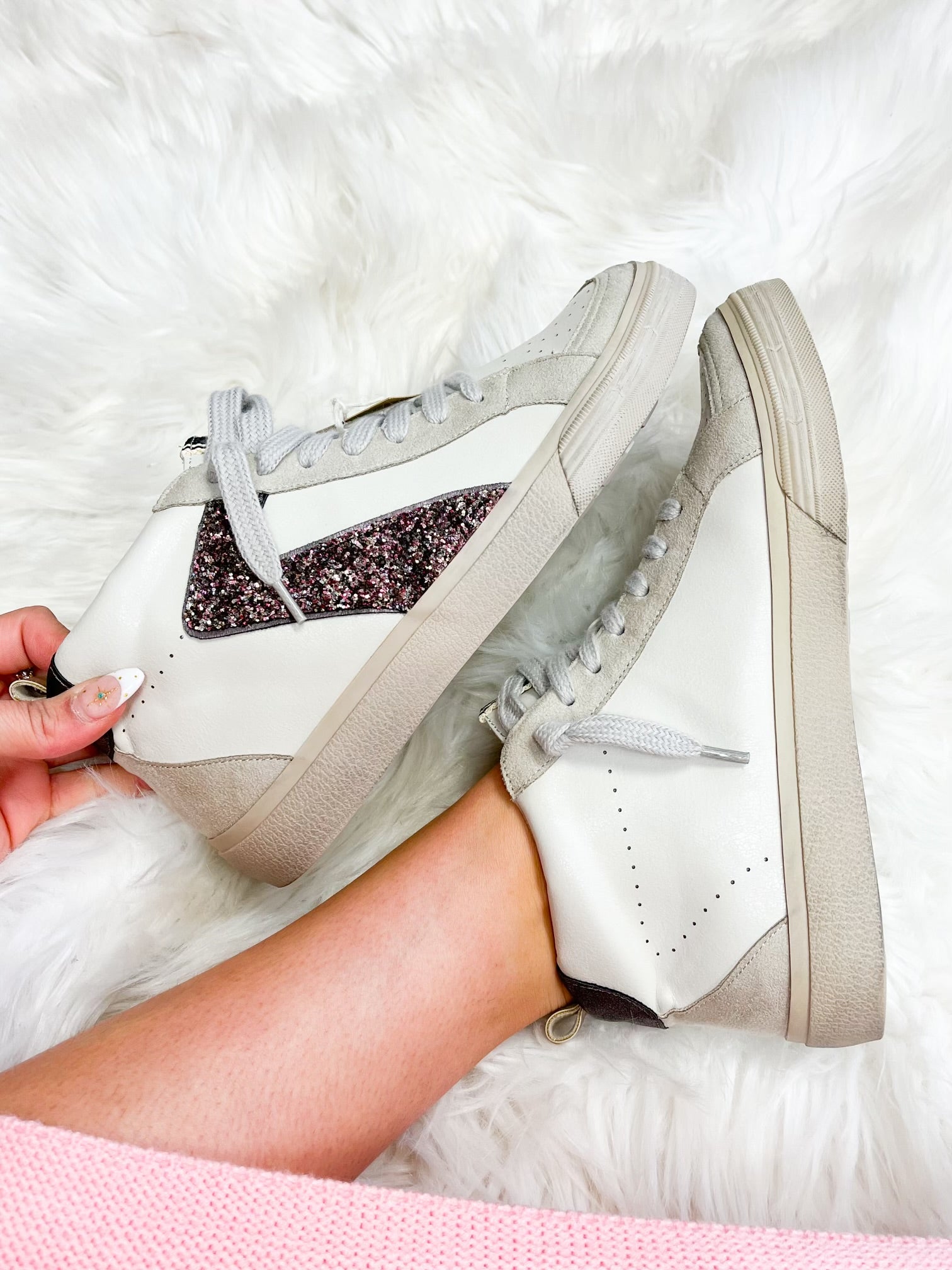 A close-up of a white sneaker with glitter on the side, held by a person against a furry white background.