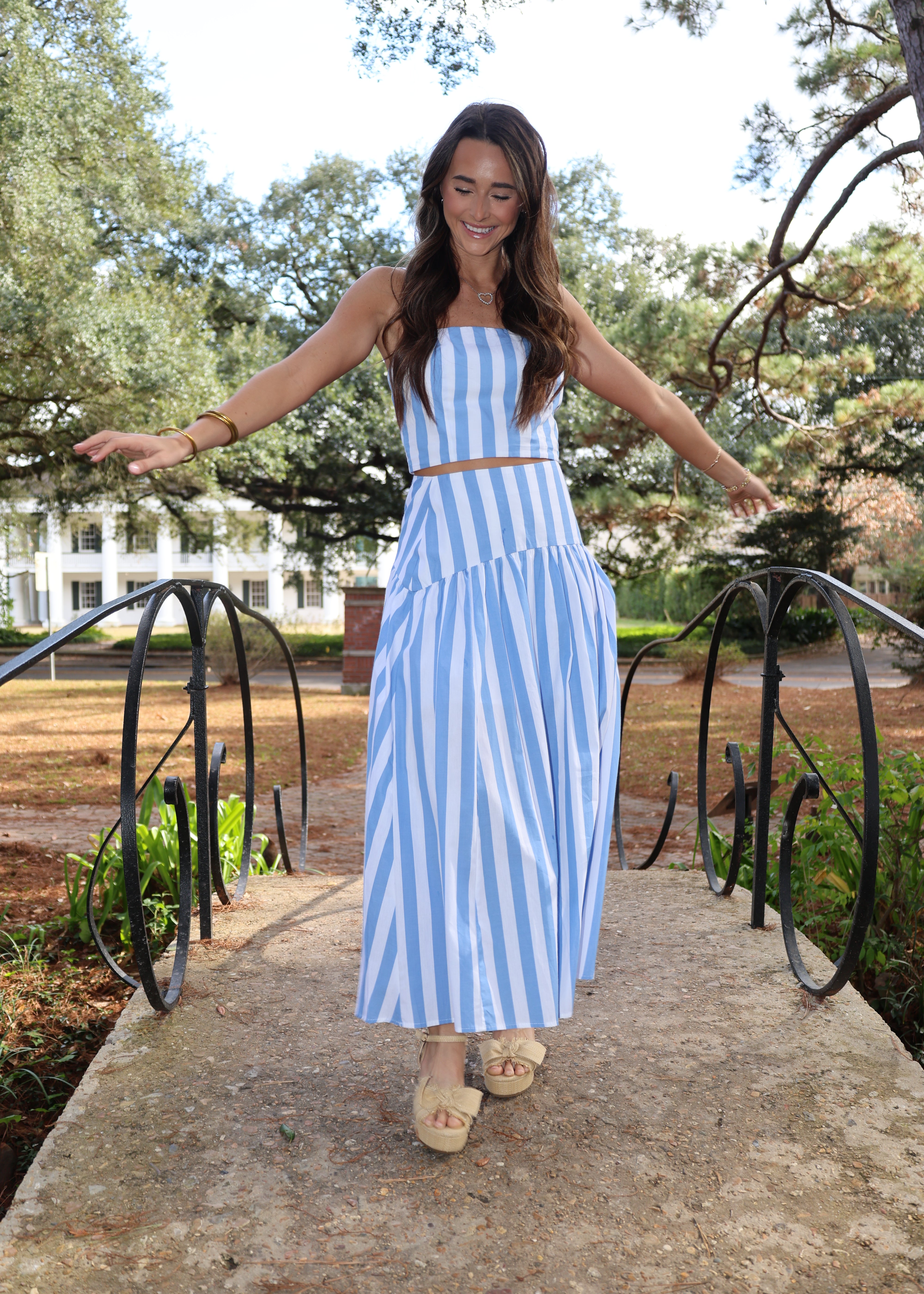 Woman in a blue and white striped dress standing outdoors with trees and a path in the background.