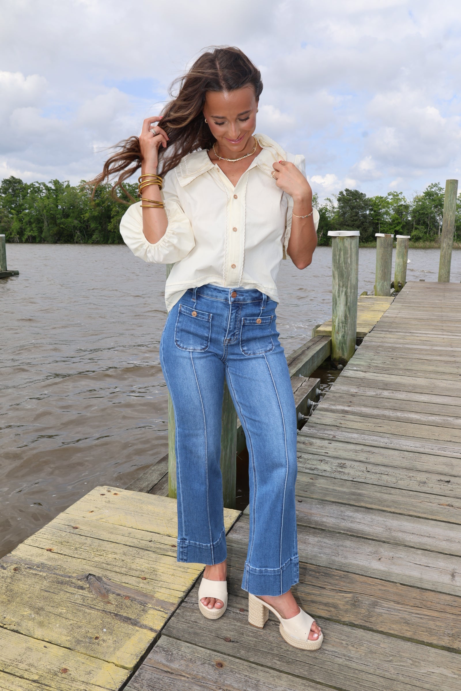 Woman in a white blouse and blue jeans standing on a wooden dock by a body of water.