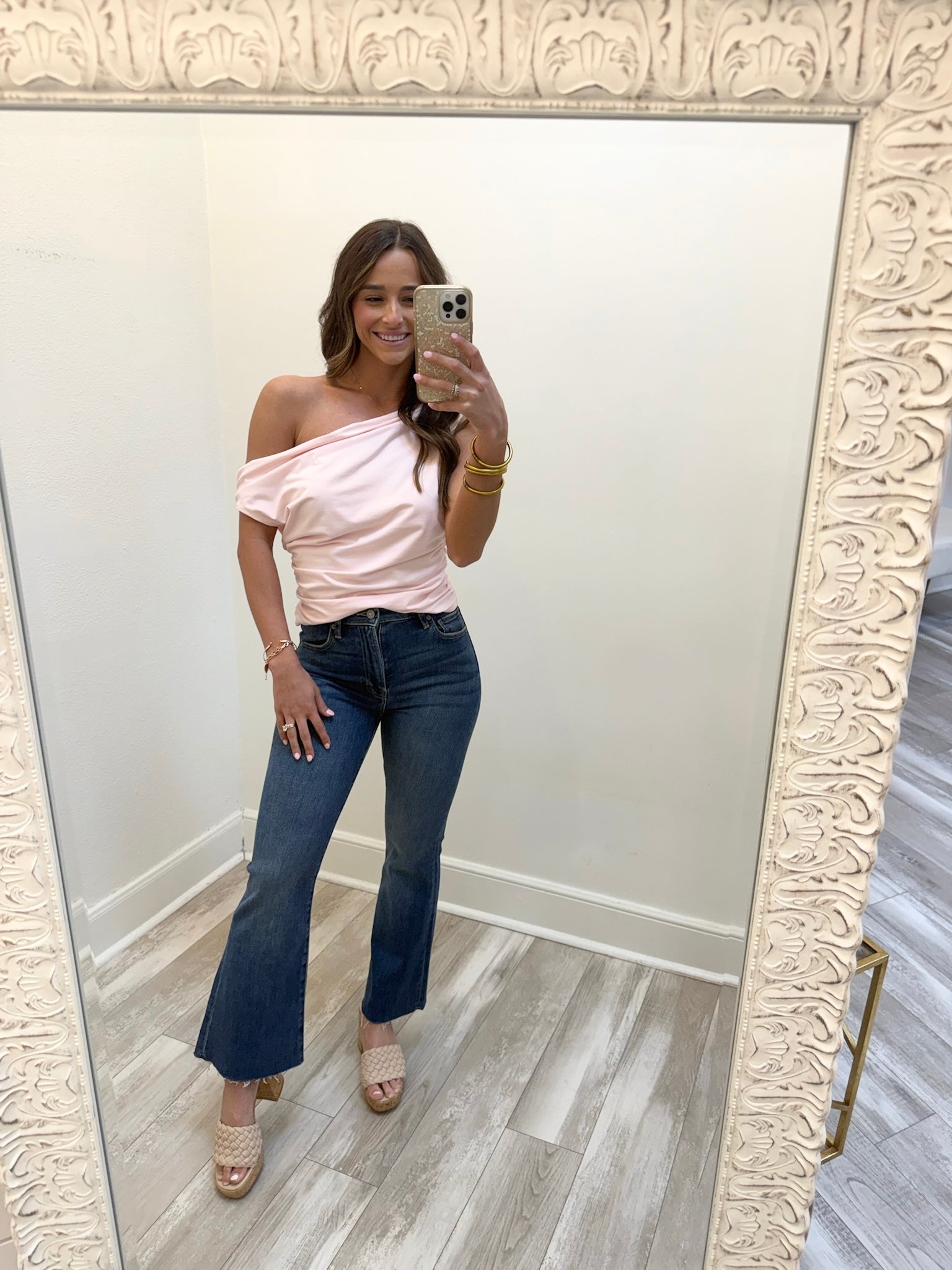Woman taking a mirror selfie wearing a pink one-shoulder top and blue jeans.