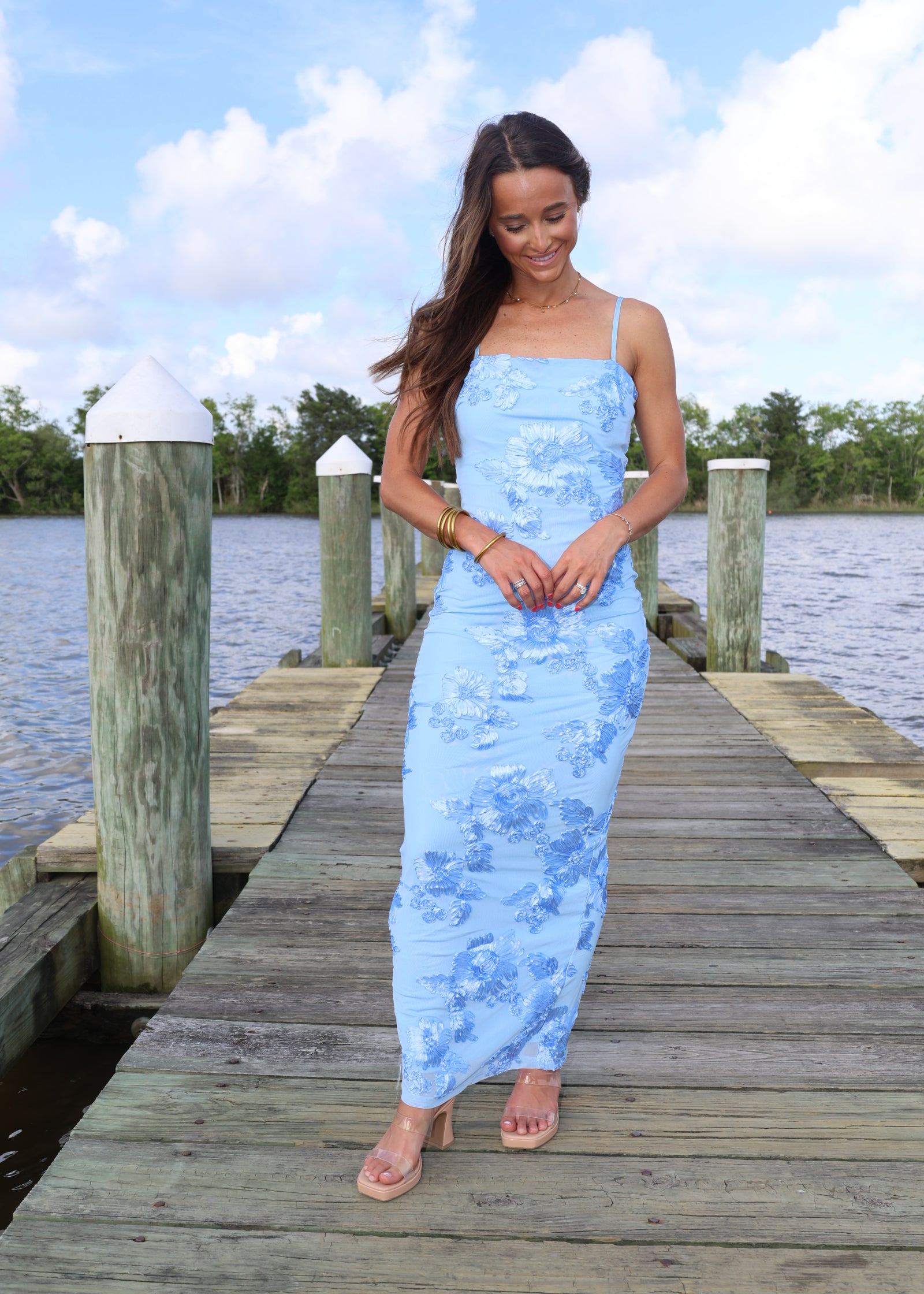 Woman in a light blue dress standing on a wooden dock by a body of water.