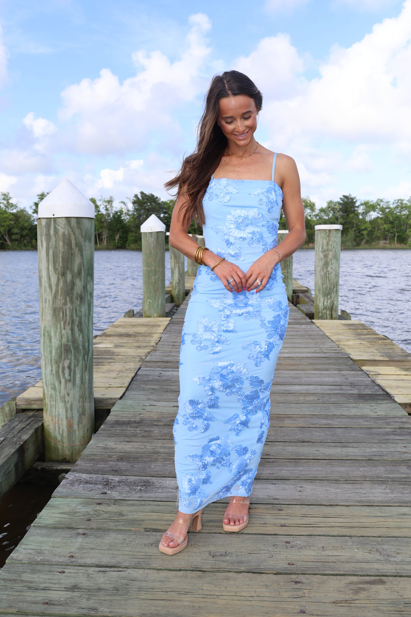 Woman in a light blue dress standing on a wooden dock by a body of water.